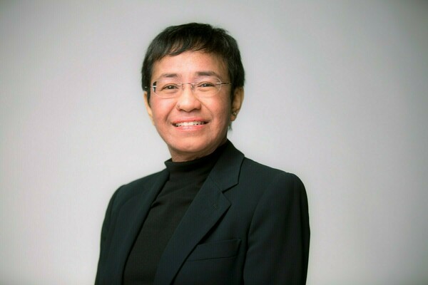 A headshot of Maria Ressa. She wears a black turtleneck and black blazer, and is smiling at the camera.